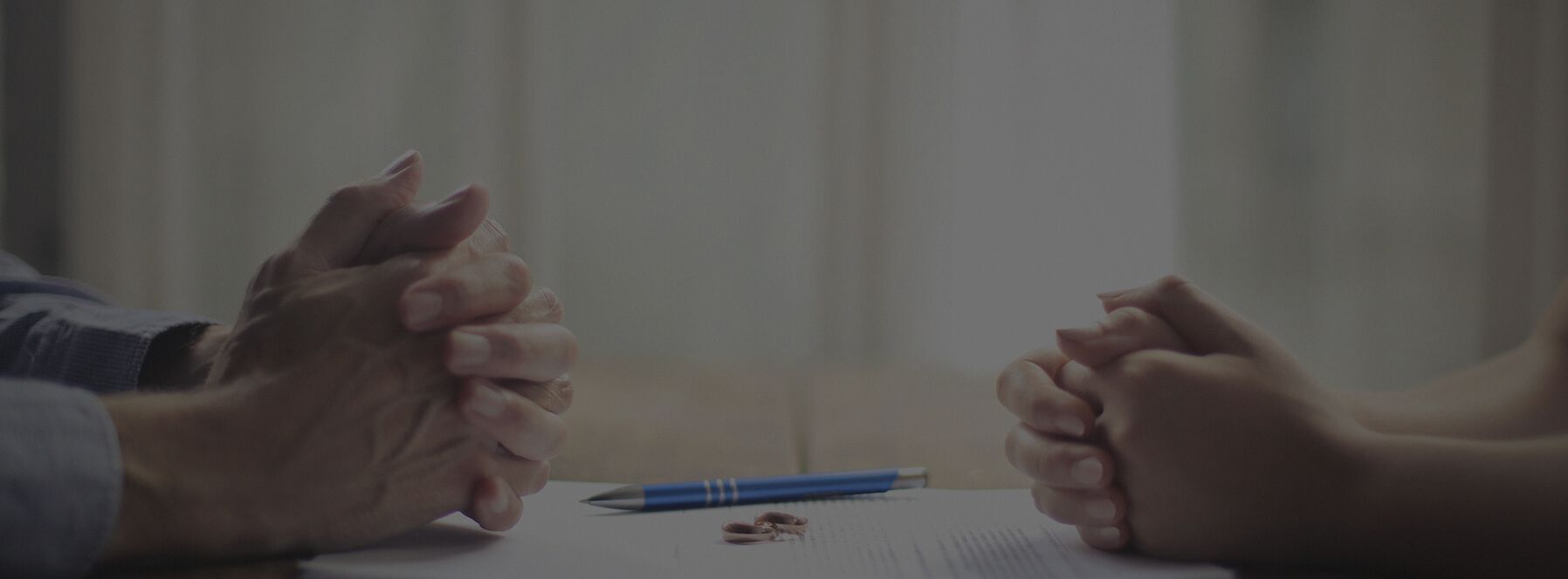 two pairs of folded hands rest on either side of a divorce agreement with wedding rings and a pen sitting on top